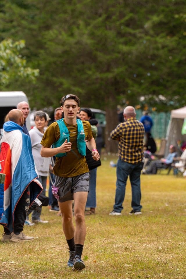 Male runner in a brown shirt and blue hydration vest jogs through a grassy field.