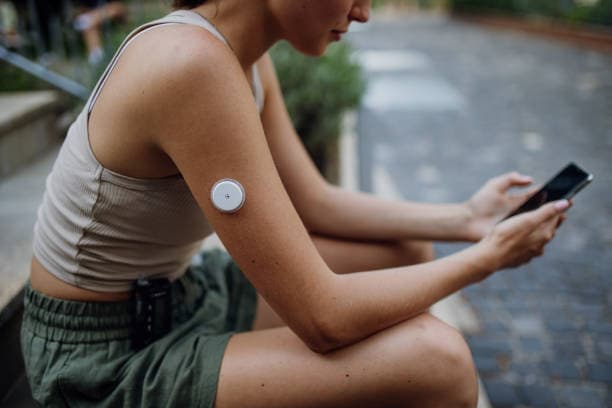 Woman sitting outdoors checking her smartphone with a continuous glucose monitor on her upper arm.
